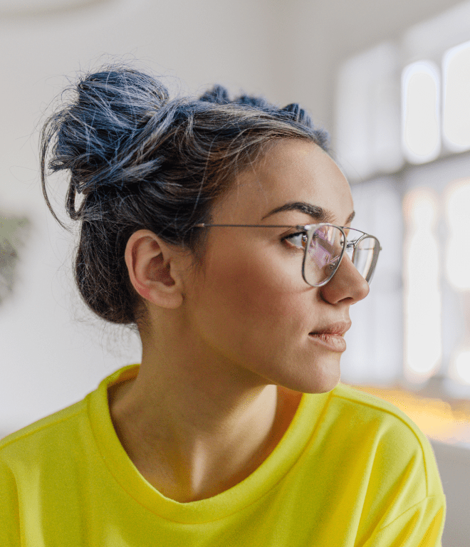 woman in a yellow shirt with blue hair pigtail buns wearing glasses looking out window.
